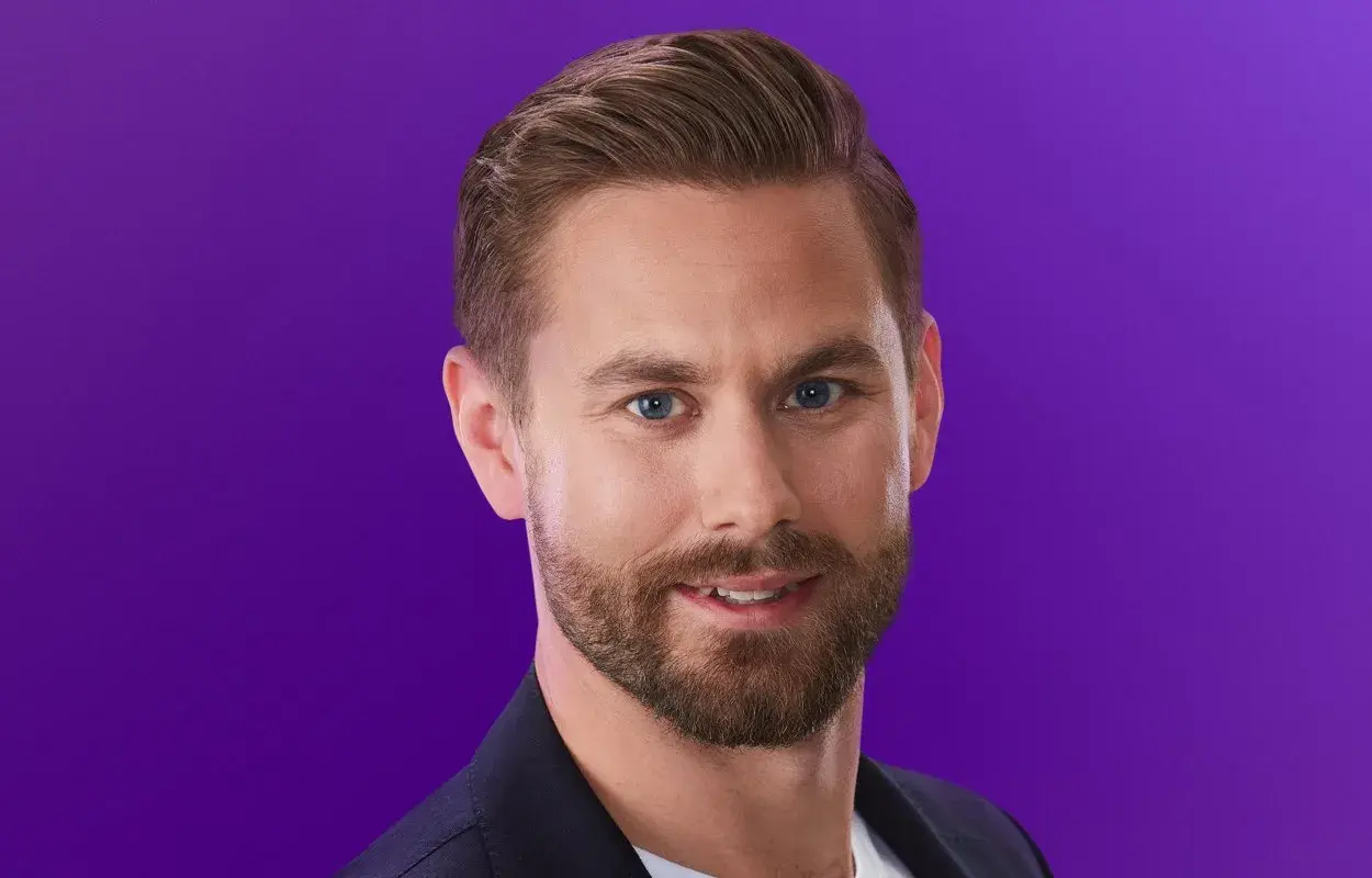 Oskar från Love is Blind, med blå ögon och skägg, ler mot kameran mot en lila bakgrund.