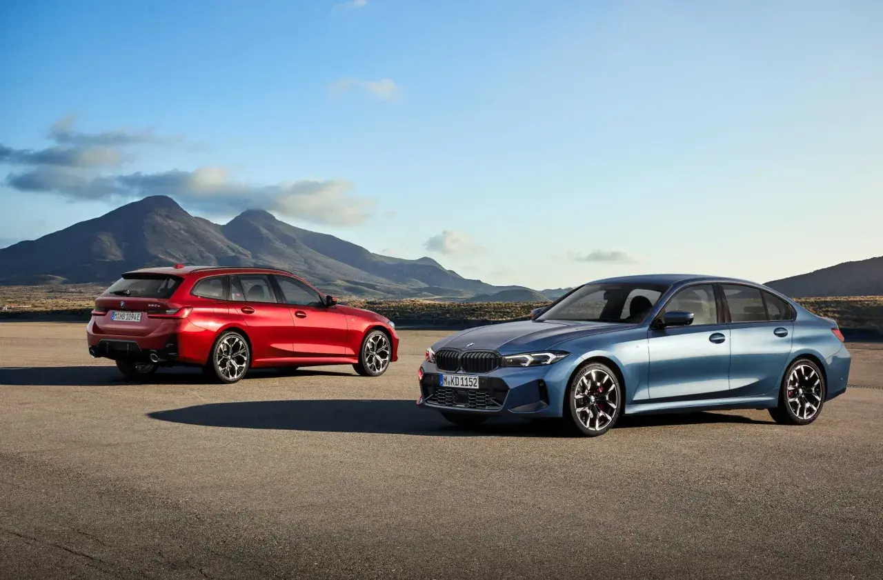 Dwa nowe BMW Serii 3 2025: niebieski sedan i czerwone kombi, stoją na pustkowiu z górami w tle.