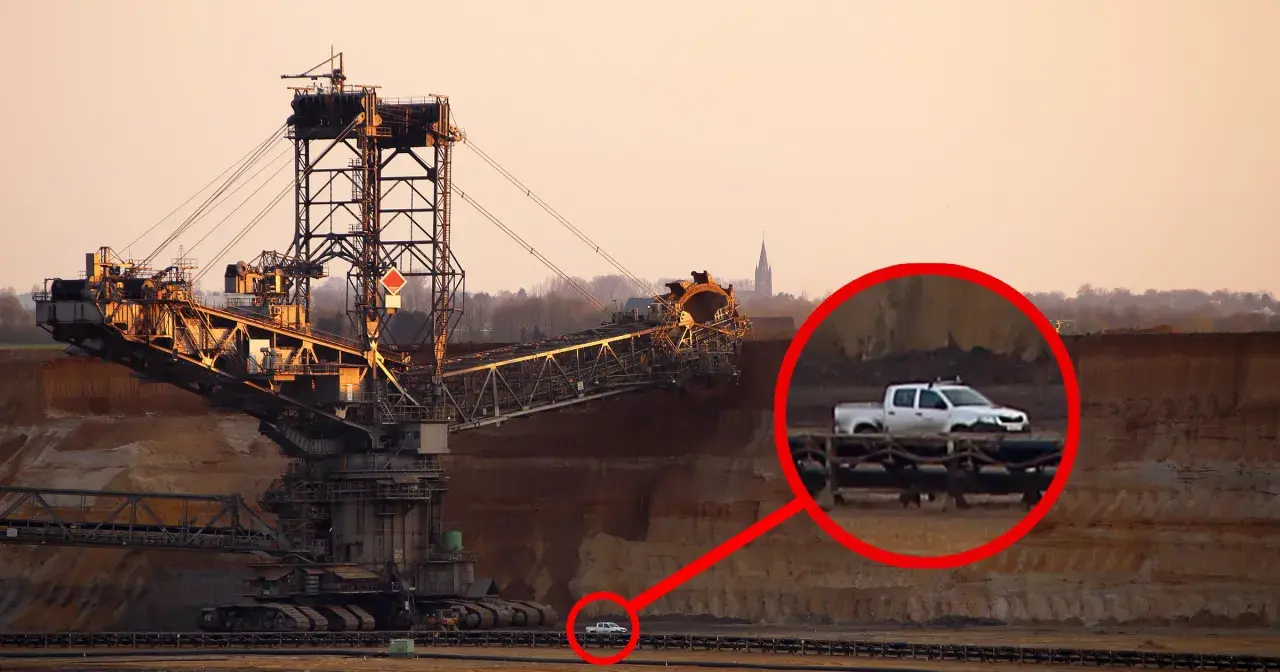 Największa koparka świata w kopalni odkrywkowej. W oddali widać mały biały pickup, który wygląda jak zabawka przy gigantycznej maszynie.