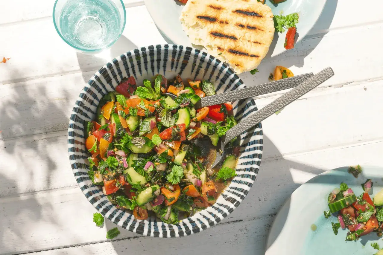 Frischer Tomaten Gurken Salat mit Kräutern, serviert in einer Schüssel mit blau-weißen Streifen. Daneben ein Glas Wasser und gegrilltes Fladenbrot.