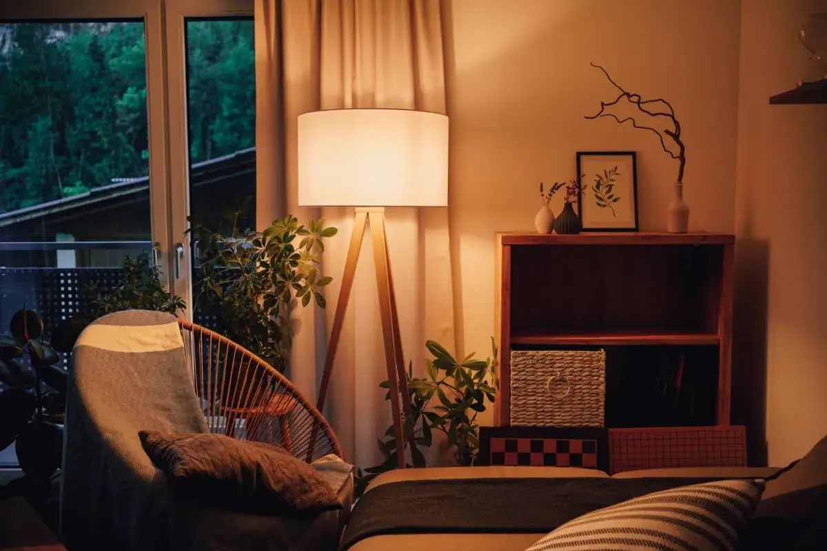 Lámpara de pie con luz blanco cálido ilumina un sillón acogedor y plantas. Estantería de madera con objetos decorativos y cestas.