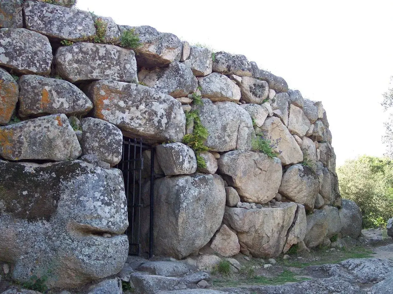 Muro imponente di un nuraghe, con un cancello in ferro che custodisce l'ingresso. Le pietre antiche, segnate dal tempo, raccontano storie millenarie.