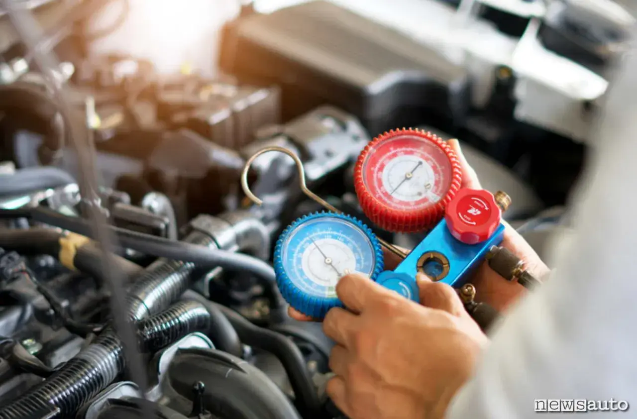 Manometro per ricaricare aria condizionata auto, con due quadranti (rosso e blu) collegati a tubi, in mano a un meccanico davanti al motore.