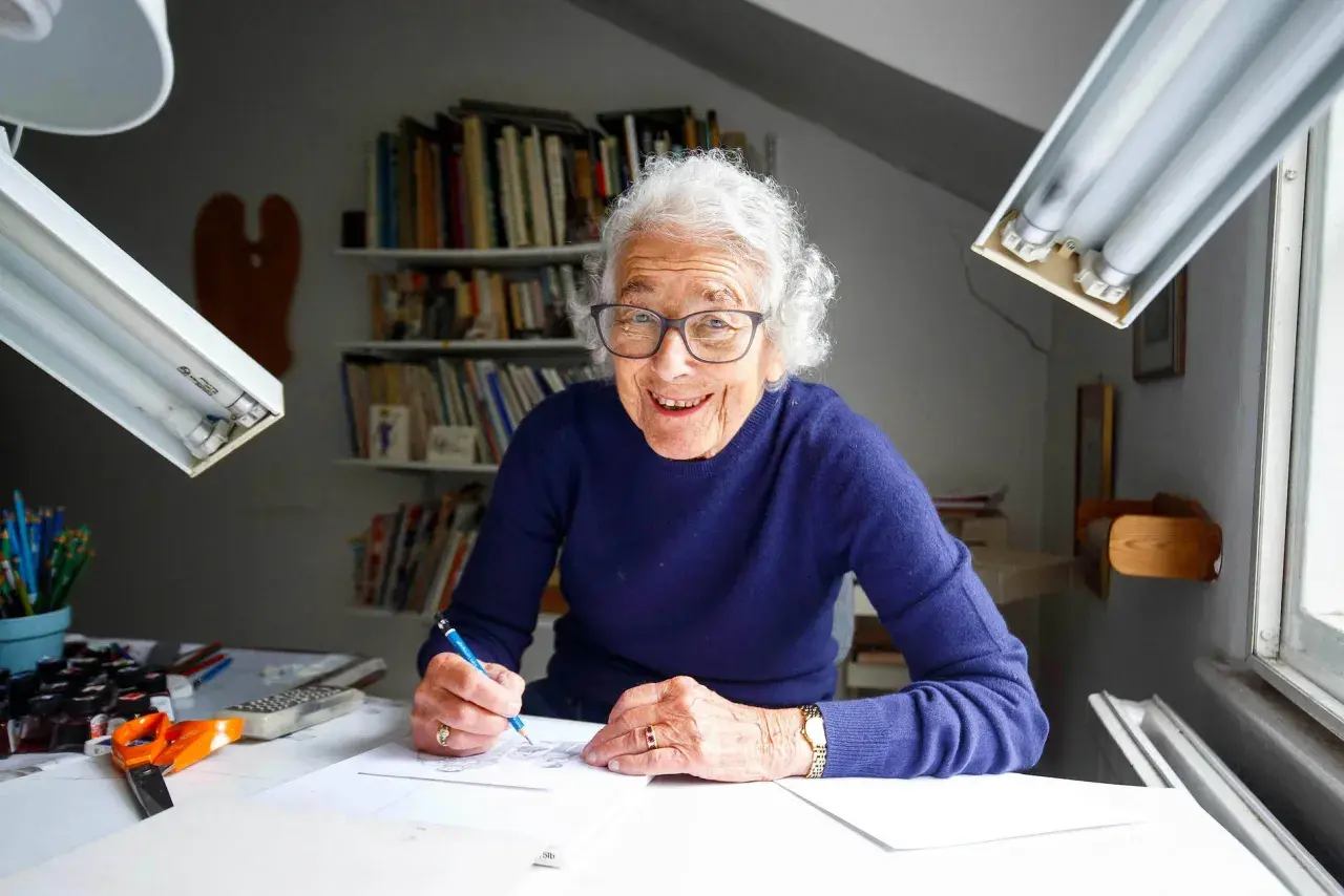 Judith Kerr, lächelnd, zeichnet an ihrem Schreibtisch. Bücherregale im Hintergrund.