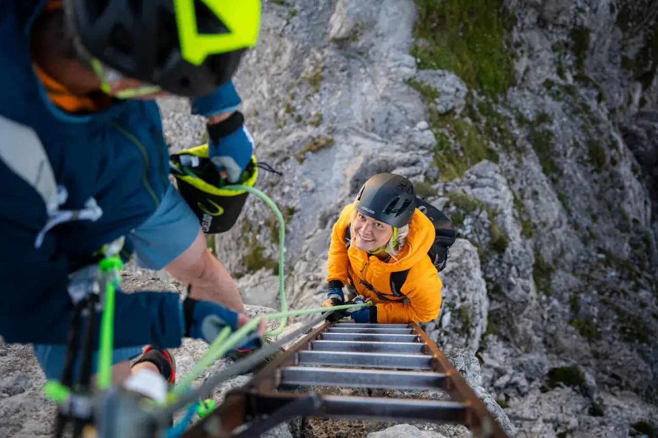 Dos personas con equipo ferrata escalan una pared rocosa, una subiendo una escalera metálica y la otra asegurándola.