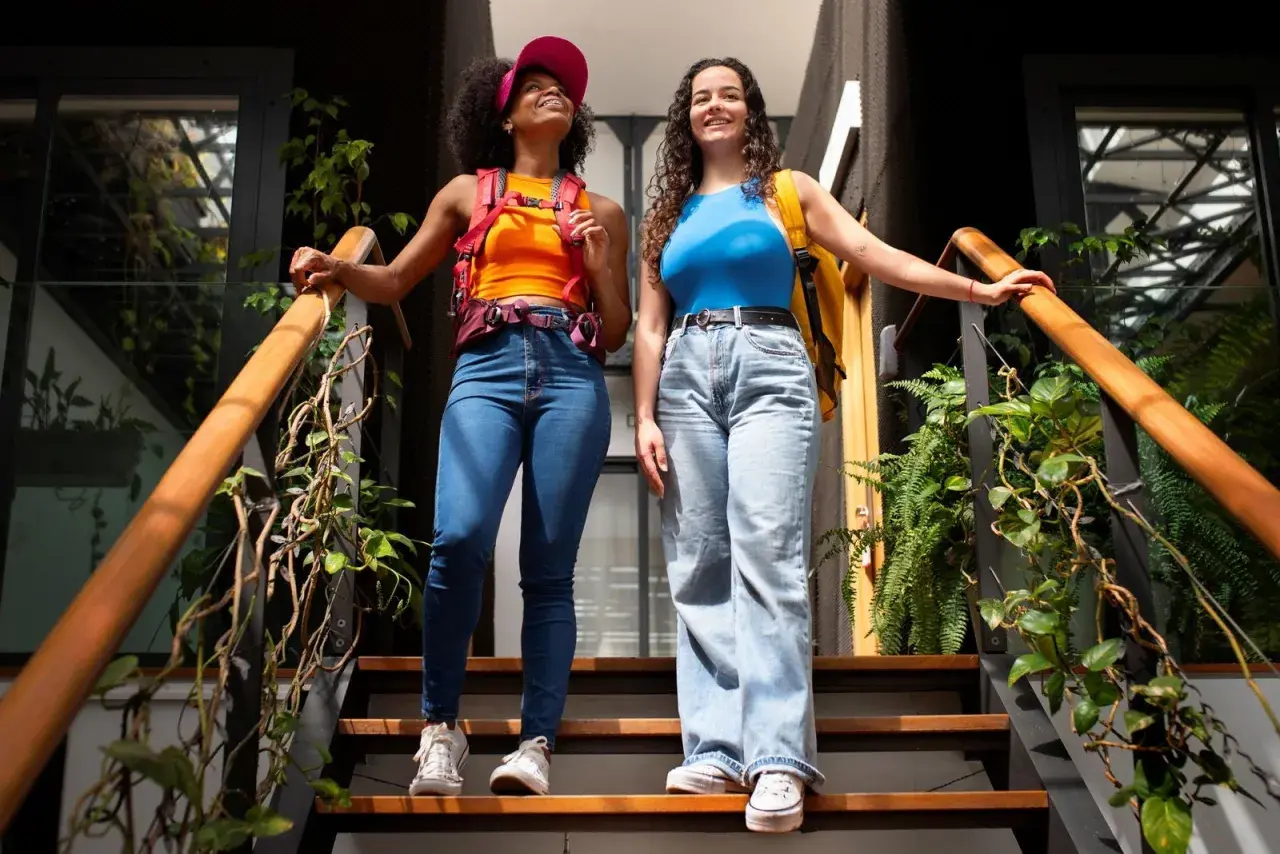 Dos amigas con estilo y2k, una con top naranja y gorra rosa, la otra con top azul, bajan unas escaleras rodeadas de plantas.