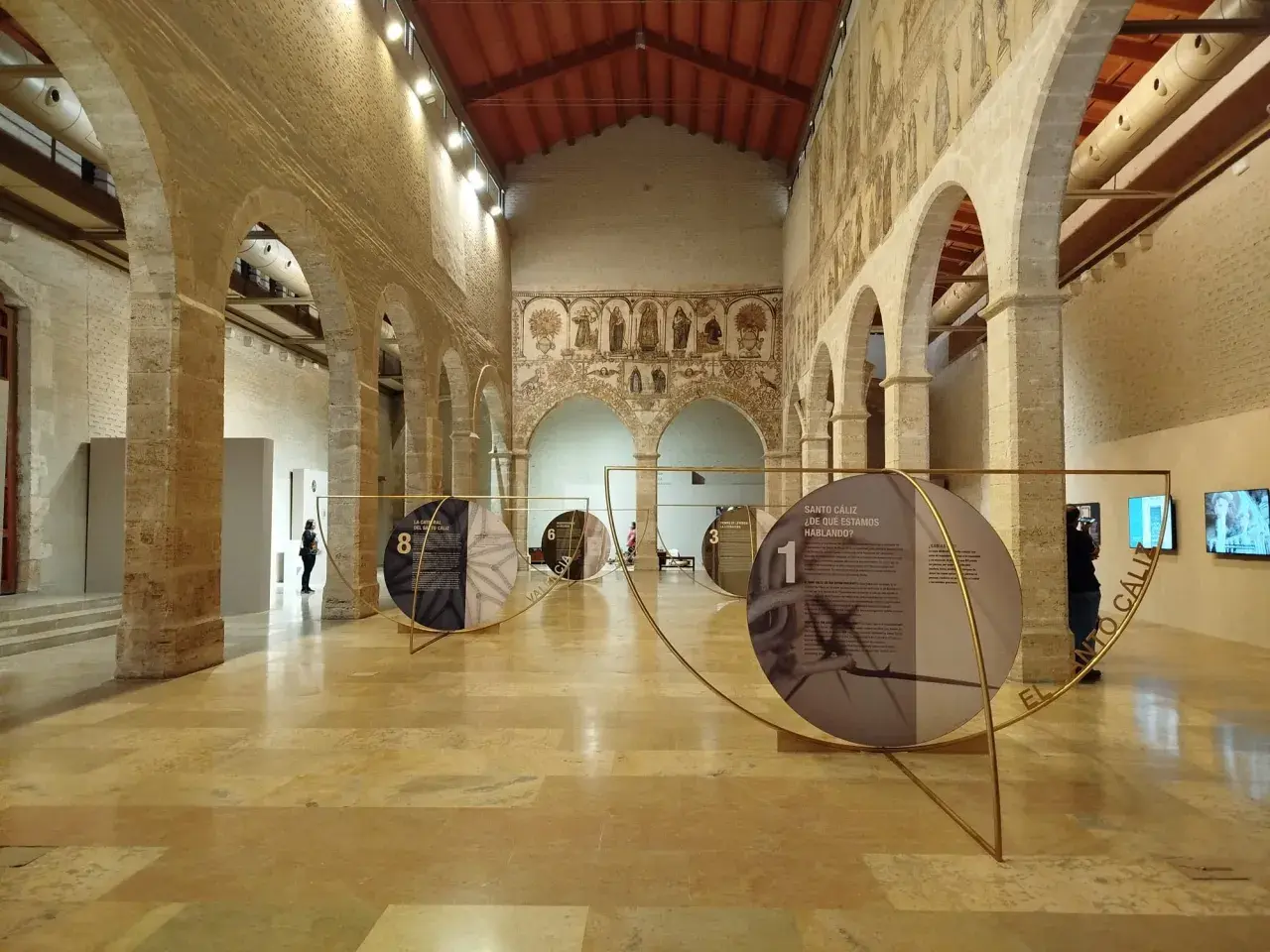 Exposición en el Almudín de Valencia, con arcos de piedra, murales antiguos y paneles circulares que narran la historia del Santo Cáliz.