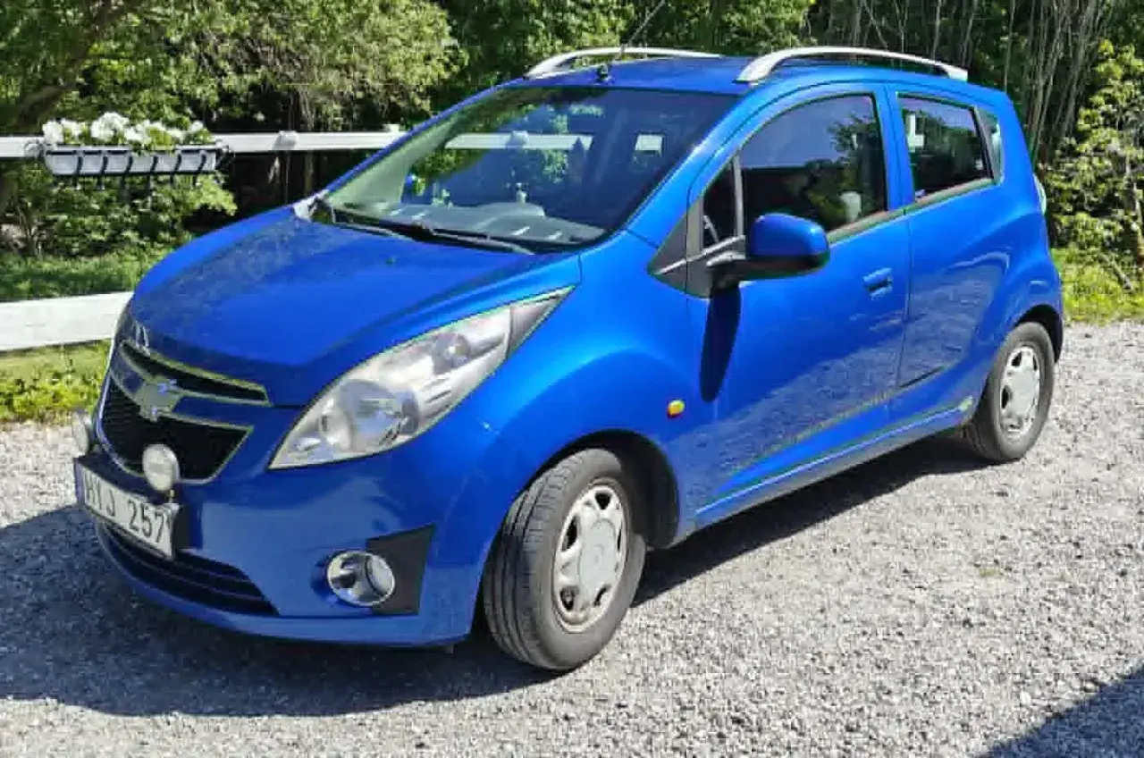 Blå Chevrolet Spark med extraljus, kanske ett problem med bilen? Parkerad på grusväg.