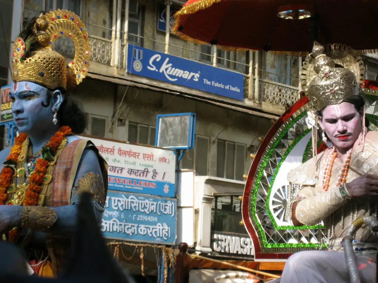 Två män i gyllene krön och smink, en med blå hud, deltar i en procession. Frågan "hur många gudar har hinduismen" kan väckas av deras utseende.