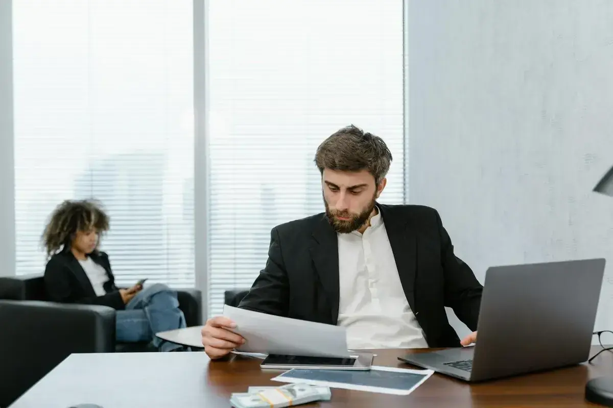 A man in a suit reviews documents, perhaps researching accelerated online MPA programs. A woman in the background is on her phone.
