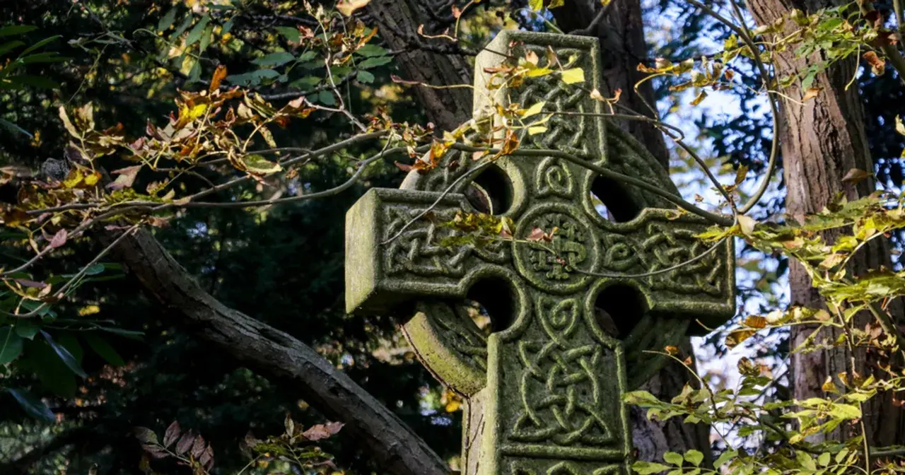 Kamienny krzyż celtycki z misternymi wzorami, otoczony jesiennymi liśćmi. Symbolizuje wieczność i połączenie życia.