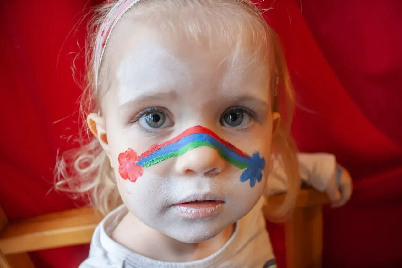 Ein kleines Mädchen mit einem Regenbogen im Gesicht und Blumen auf den Wangen. Kinderschminken einfach gemacht!