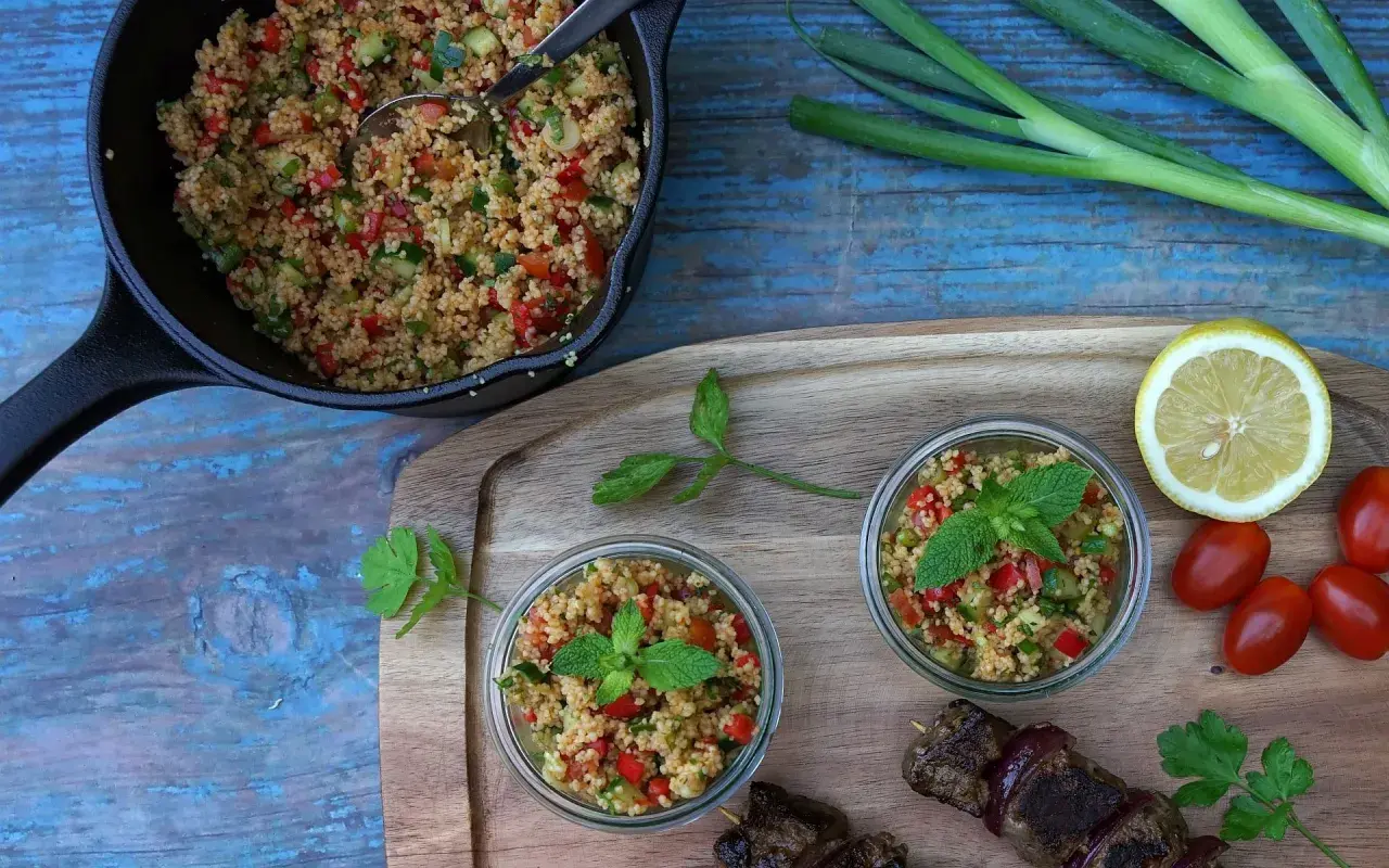 Frischer Couscous-Salat mit Tomaten, Gurken und Minze, serviert in Gläsern und einer Pfanne, daneben ein Spieß mit Fleisch.