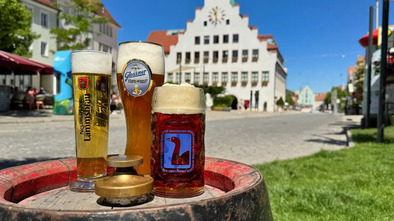 Drei Gläser Bier, darunter ein helles und ein dunkles, stehen auf einem Fass. Im Hintergrund eine malerische Stadtkulisse mit einem historischen Gebäude. Ein perfekter Genuss von italienischem Bier.