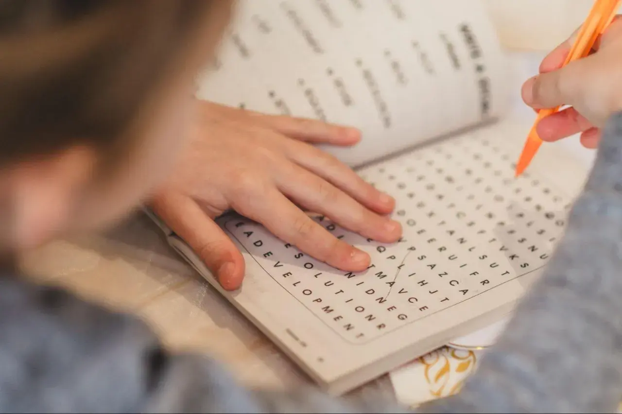 Mano de niño resolviendo una sopa de letras para la memoria con un lápiz naranja.