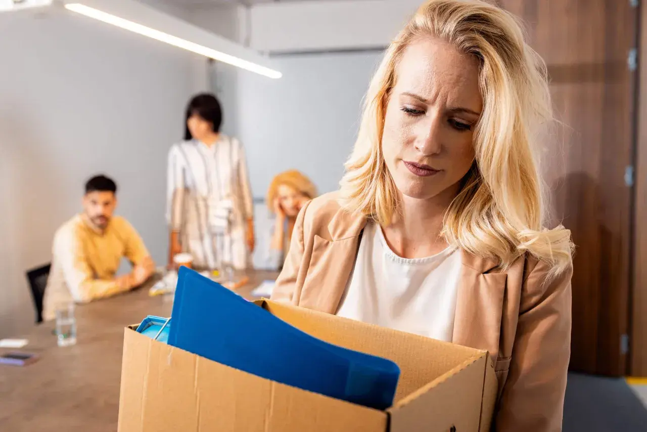 Femme blonde triste portant une boîte de ses affaires, se demandant si l'on peut licencier sans motif. Des collègues flous en arrière-plan.
