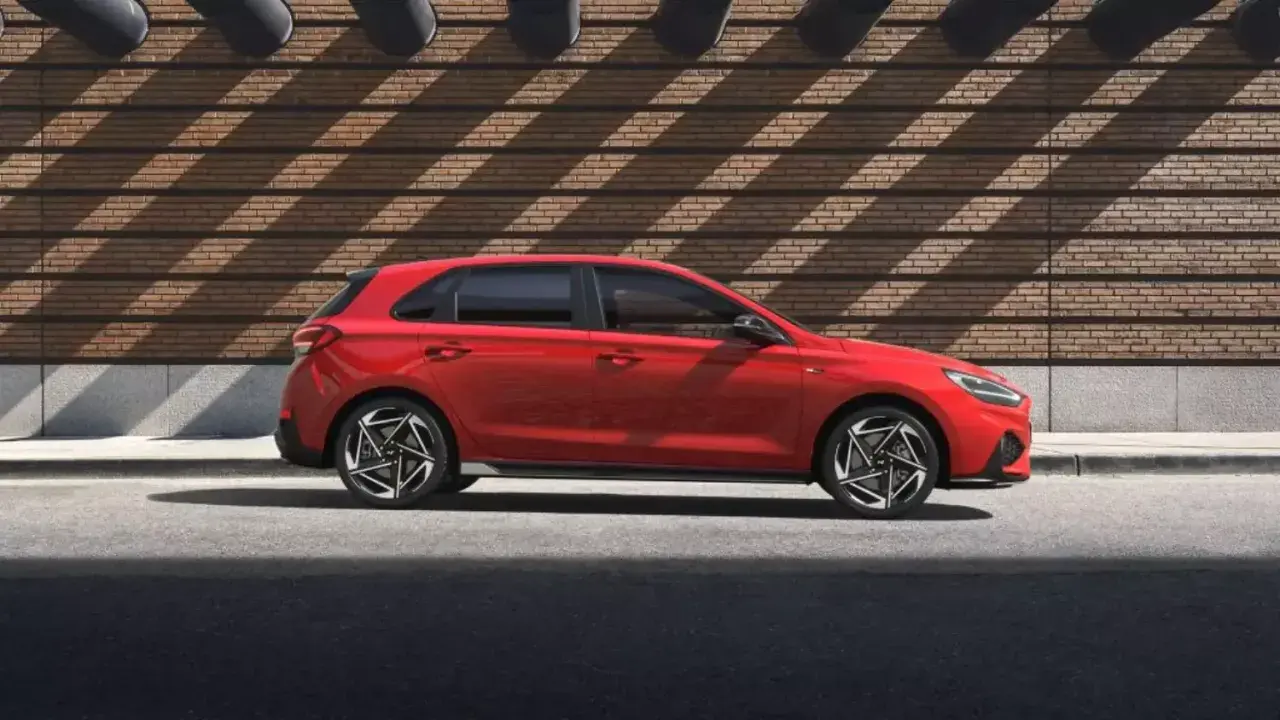Czerwony Hyundai i30 Hatchback zaparkowany na tle ceglanej ściany z geometrycznymi cieniami.