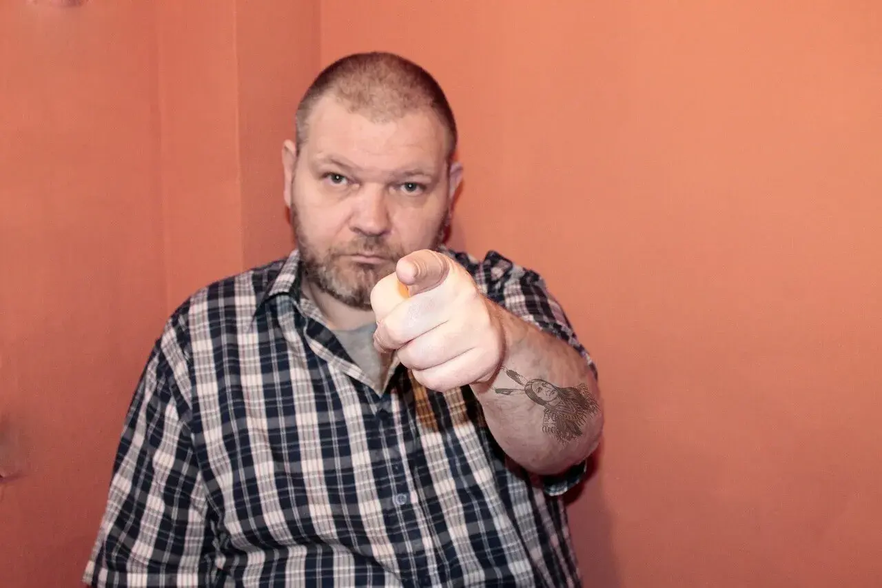 Mężczyzna w kratę wskazuje palcem, na ramieniu tatuaż z Indianinem. "234 kk" - czy to jego numer?
