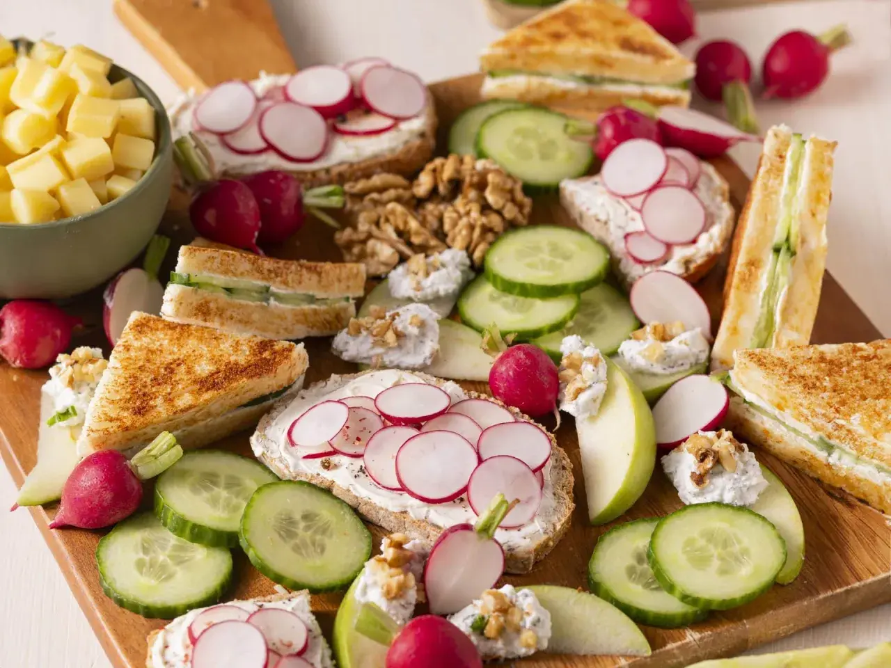 Un plateau d'apéro dinatoire plancha : toasts au fromage frais et radis, mini sandwichs concombre, dés de fromage, noix et quartiers de pomme.