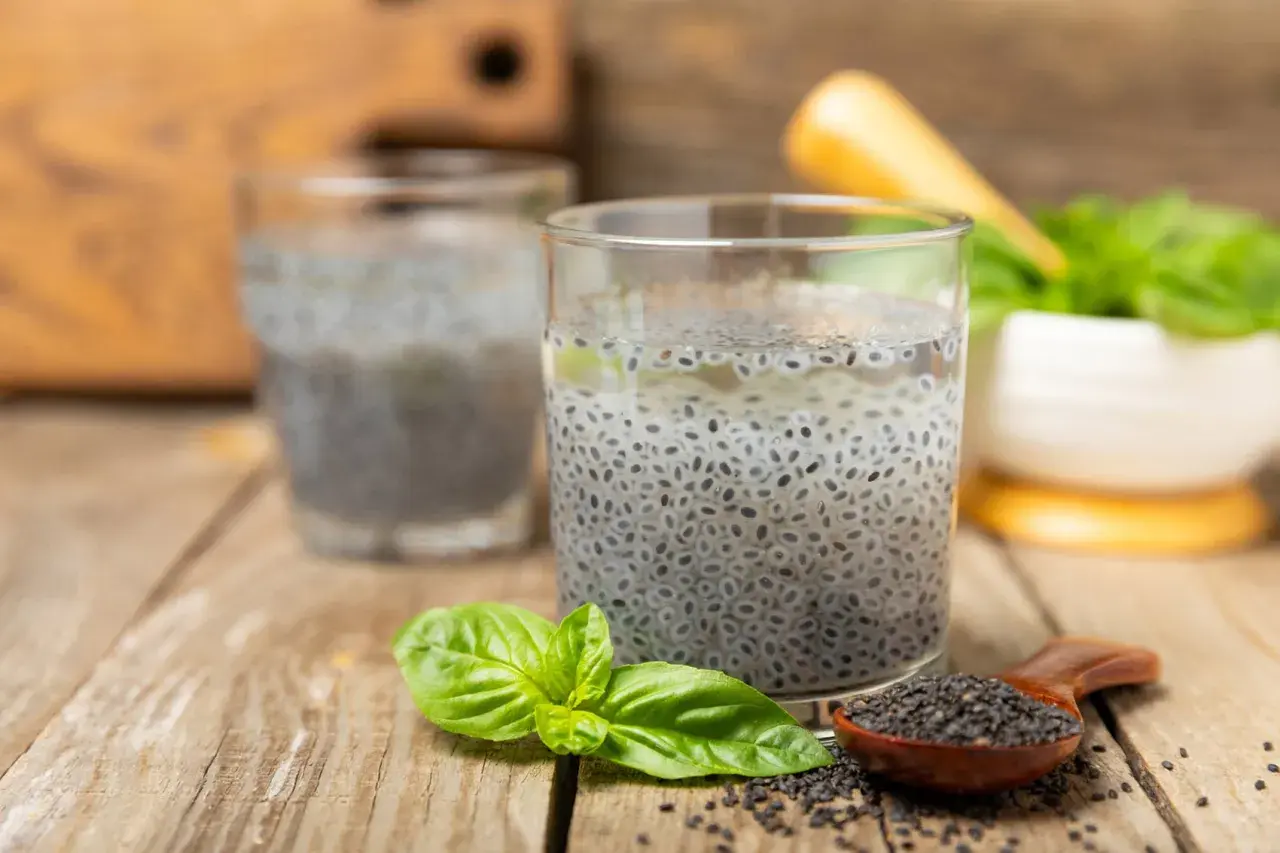 Due bicchieri con semi di basilico in acqua, un cucchiaino di semi e foglie fresche su un tavolo di legno.