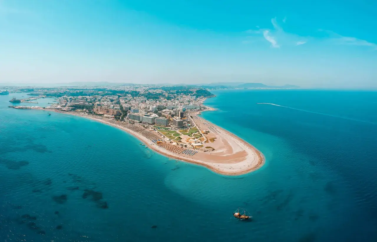 Luftfoto över Rhodos kustlinje med en sandstrand, hotell och turkost vatten. En segelbåt syns i vattnet, perfekt för Rhodos aktiviteter.