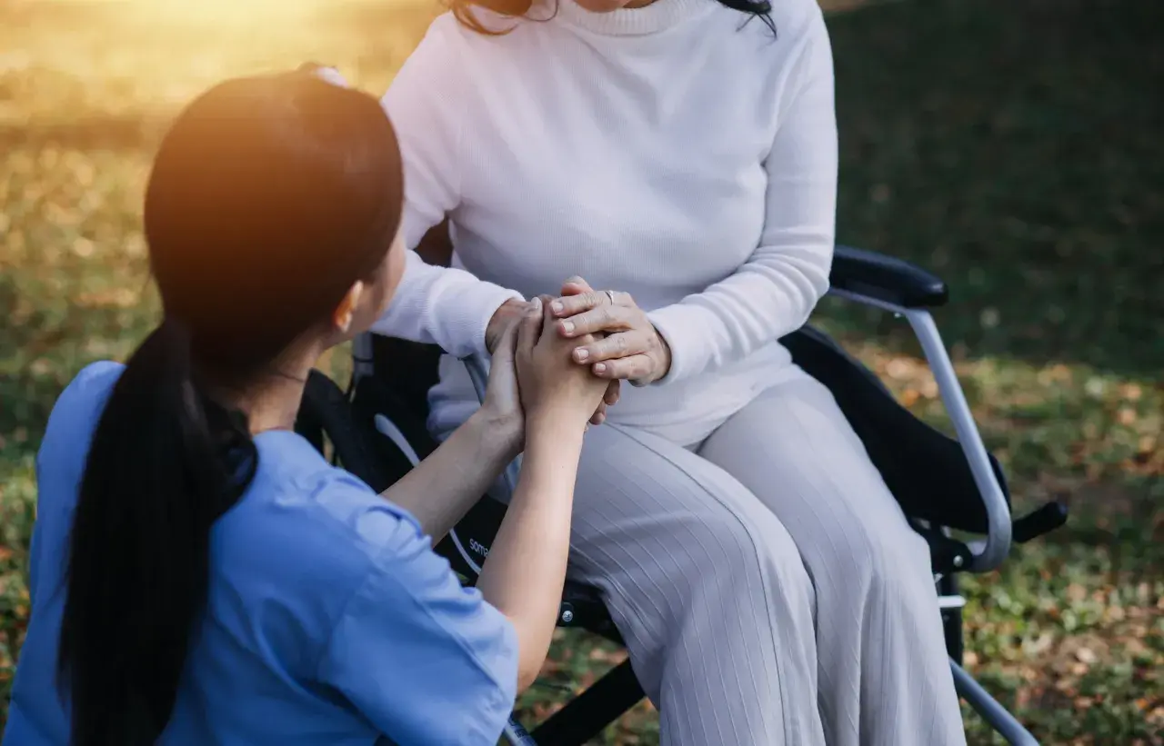 Un infermiere tiene le mani di una paziente su una sedia a rotelle, un gesto di conforto e cura. L'età non conta, l'assistenza è l'essenza.