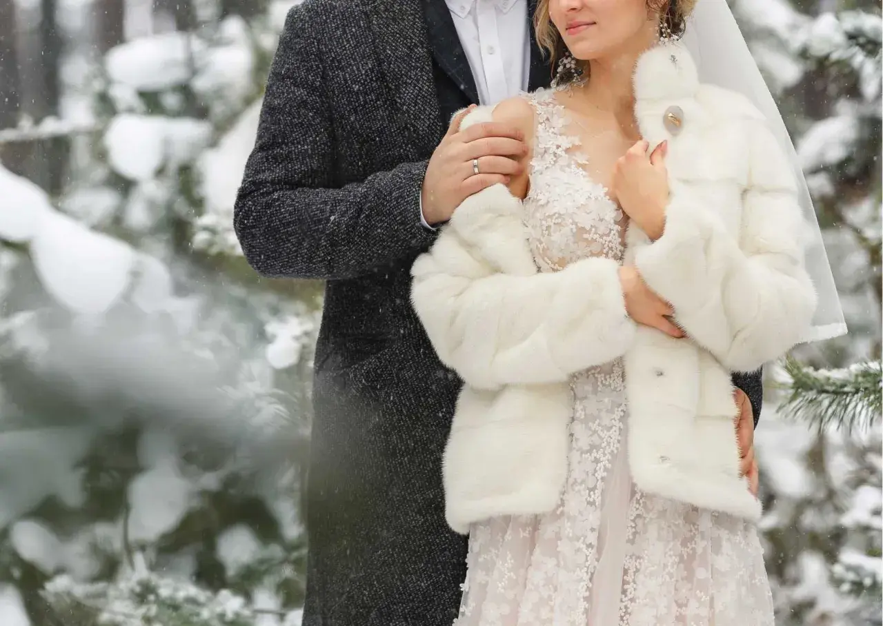 Para młoda w zimowej scenerii. Ona w białym futerku i koronkowej sukni, on w eleganckim płaszczu. Ich ślub zimą wygląda bajkowo.