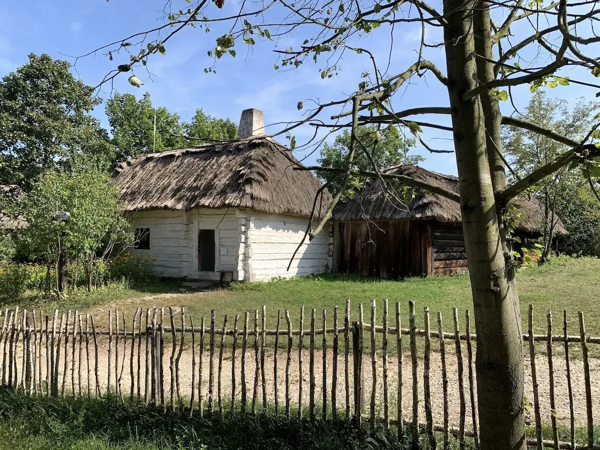Drewniane chaty kryte strzechą w skansenie, przed nimi płotek z gałęzi.