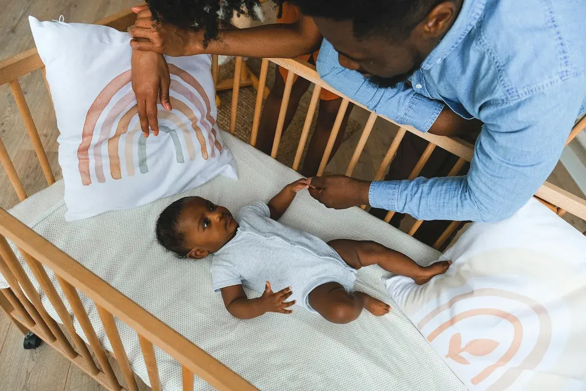 Parents hold their baby's hand in a crib with a breathable mattress, a safe sleep environment.