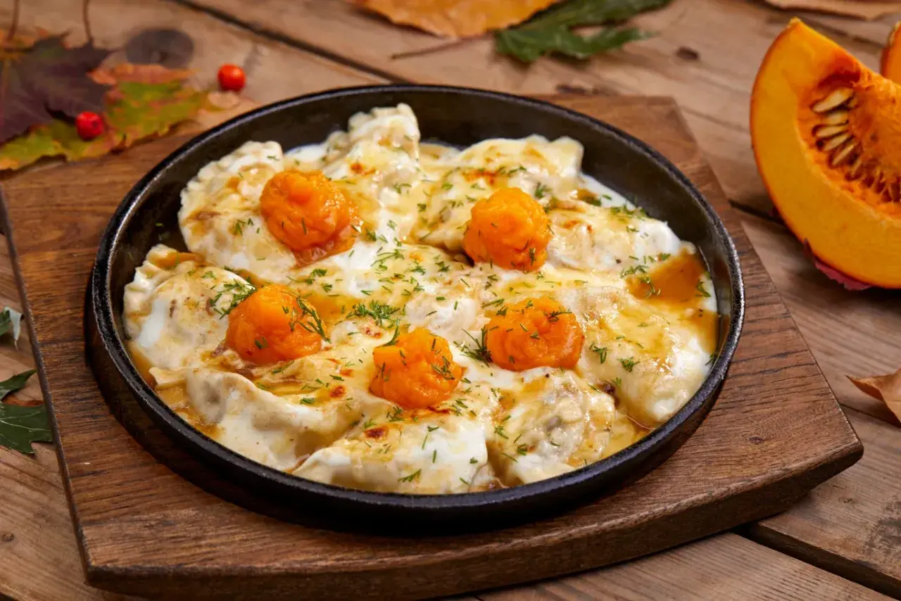 Pierożki z dynią w sosie, posypane koperkiem, podane w żeliwnej patelni na drewnianym blacie. Obok kawałek dyni.
