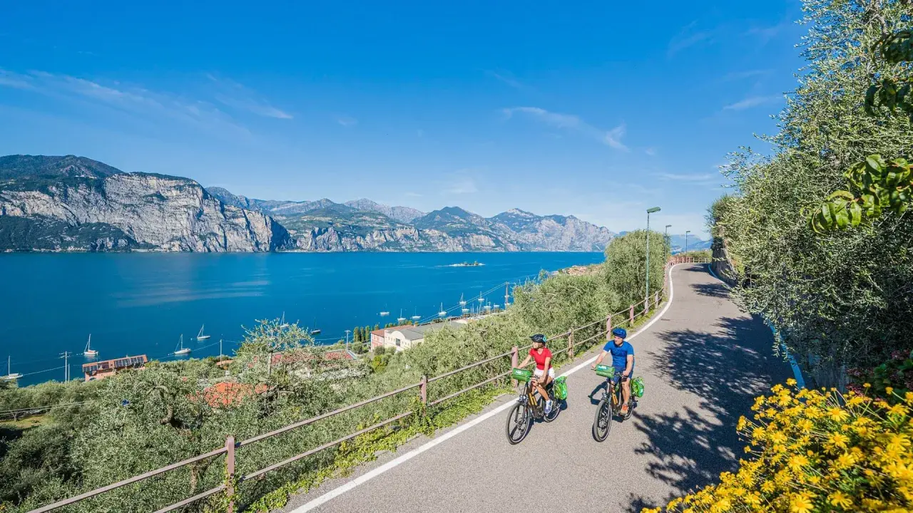 Two people enjoy biking along a scenic road in Italy, with a stunning blue lake and mountains in the background.