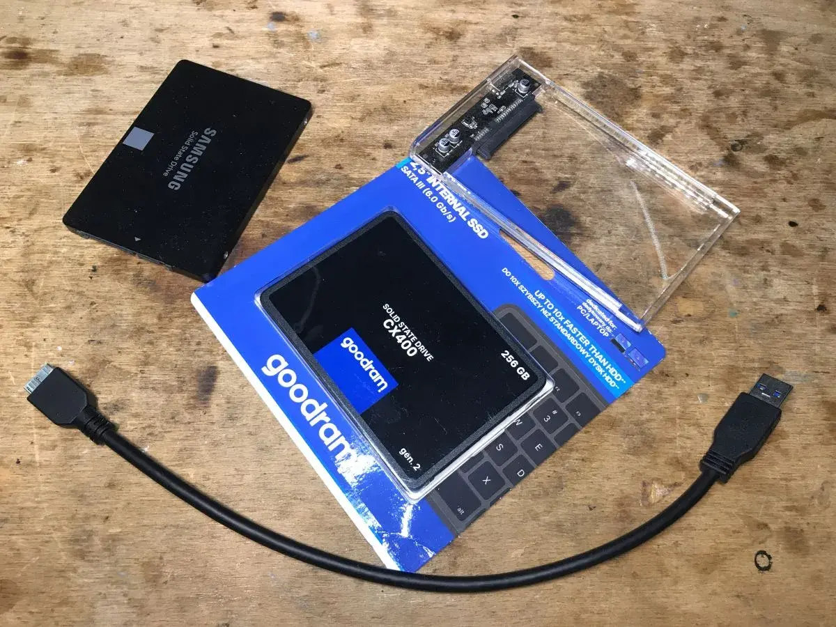 Dwa dyski SSD, jeden Samsung, drugi Goodram CX400 256 GB, obok obudowa na dysk i kable SATA.