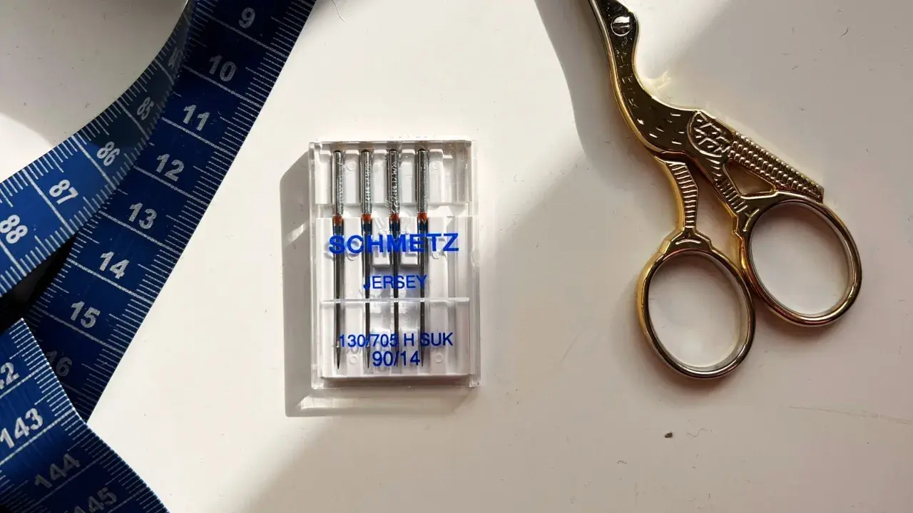 Schmetz Jersey needles, size 90/14, are ideal for stretch fabrics. A blue measuring tape and gold scissors complete the sewing setup.