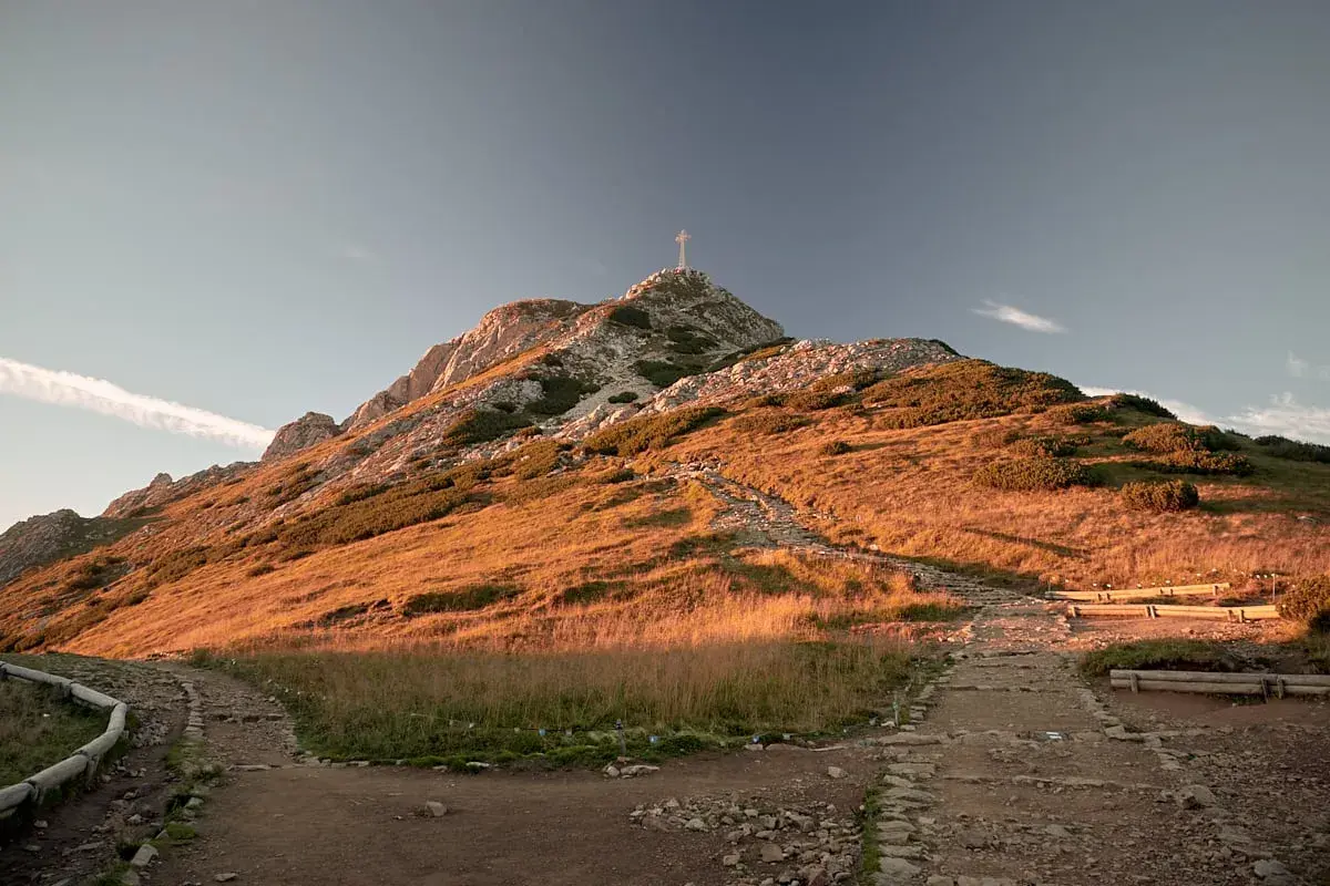 Giewont z krzyżem na szczycie, oświetlony zachodzącym słońcem. Ścieżka prowadzi w górę, a w oddali widać ławki dla turystów.