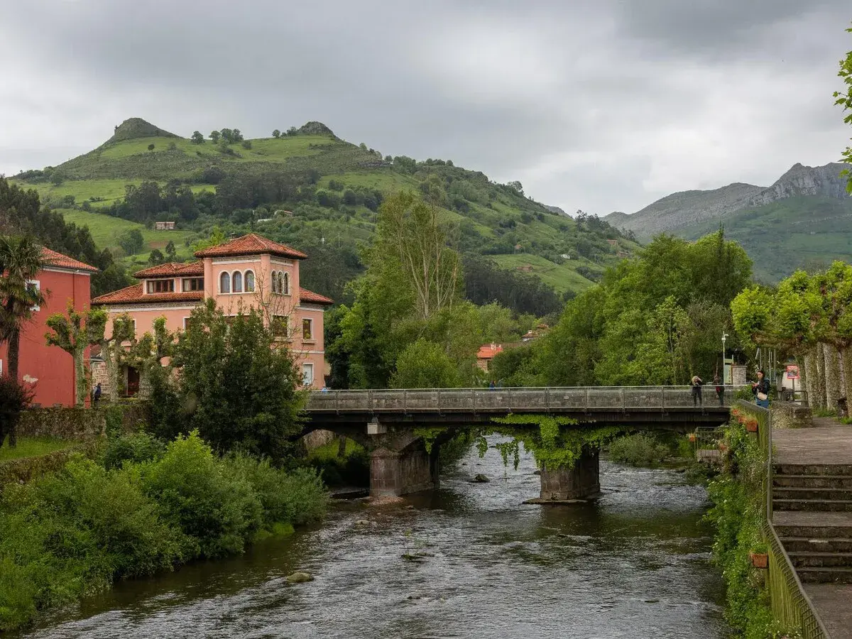Un puente cruza un río, conectando pueblos bonitos cerca de Liérganes. Edificios rosados y verdes colinas completan la escena.
