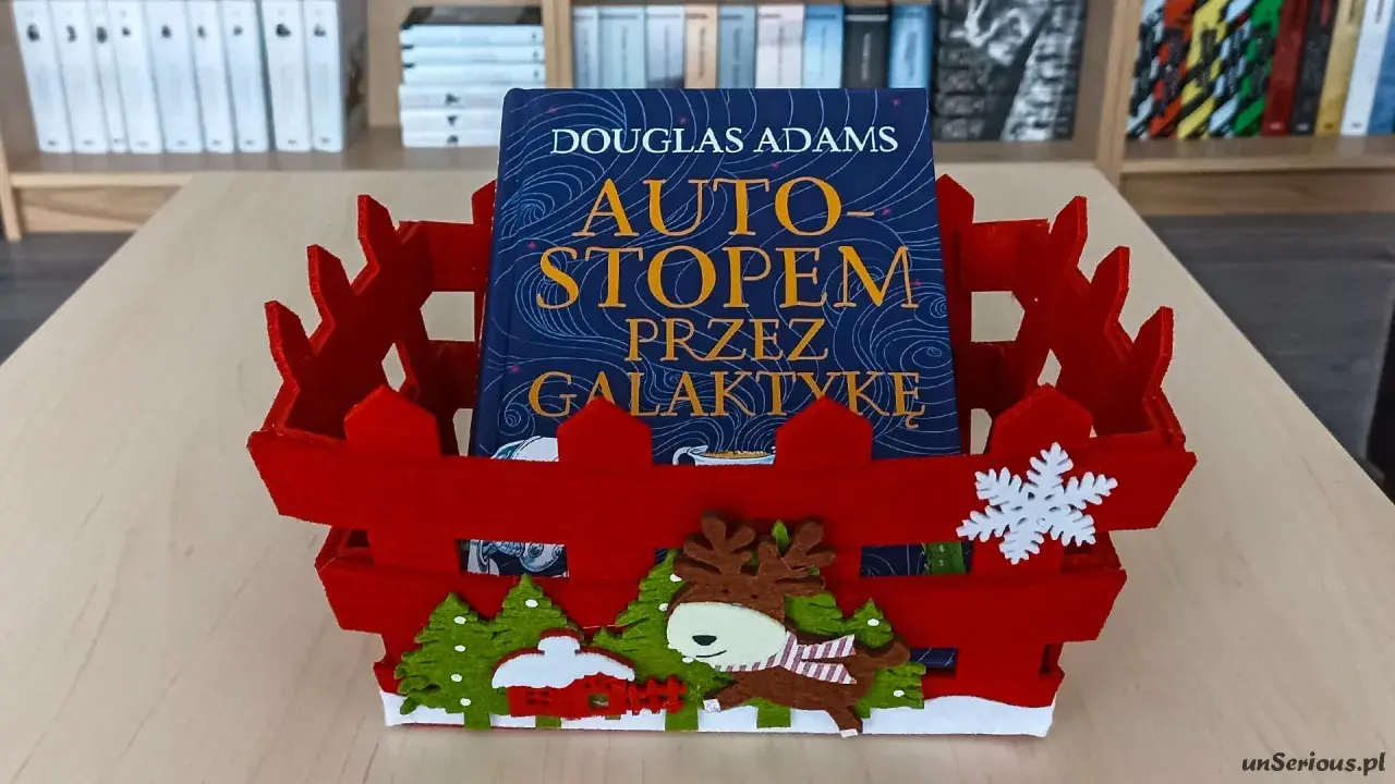 Książka "Autostopem przez galaktykę" Douglasa Adamsa w czerwznym, świątecznym koszyku z reniferem i choinkami.