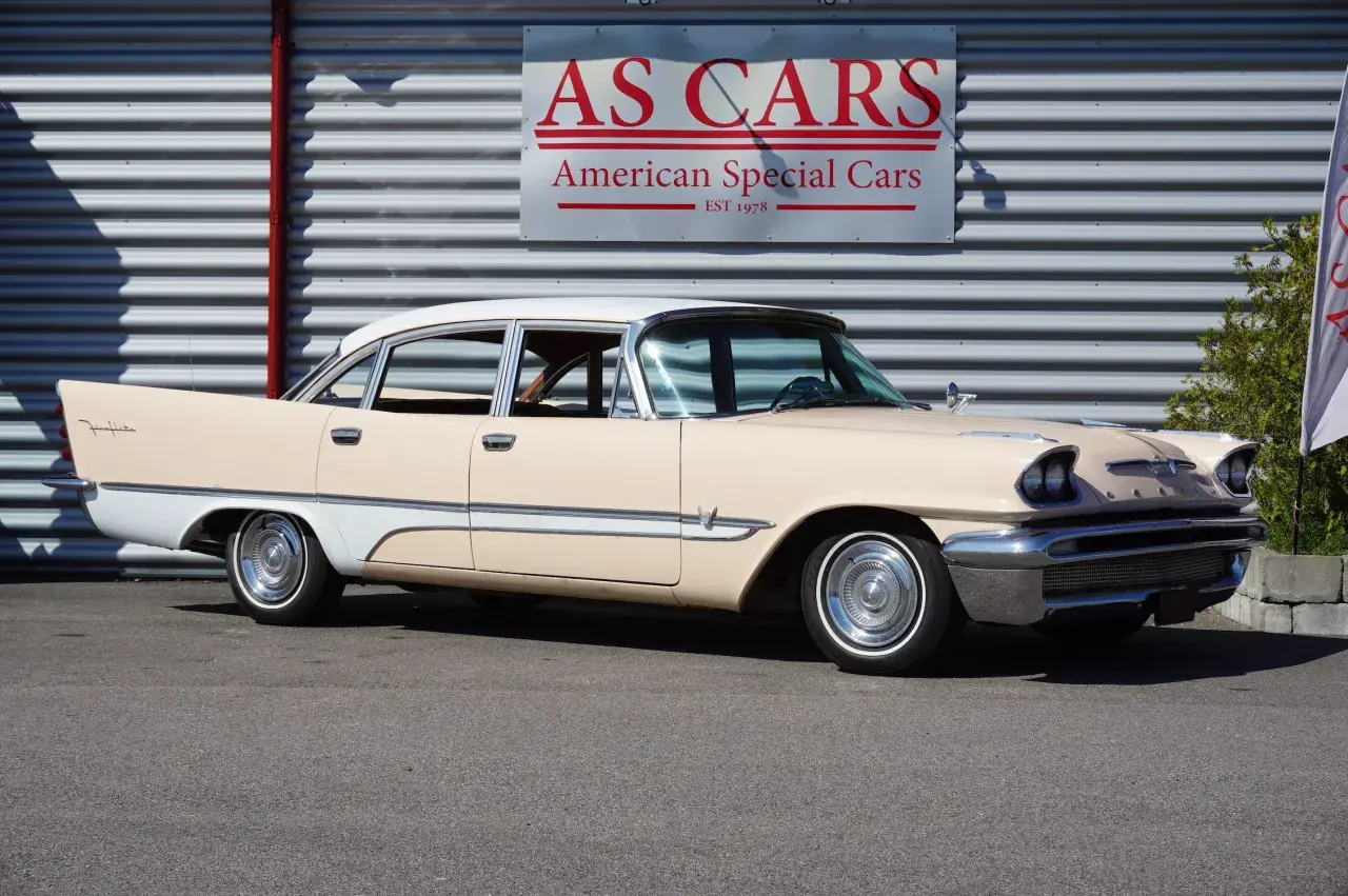En krämvit och ljusbeige De Soto från 1950-talet står parkerad framför en byggnad med skylten "AS CARS".
