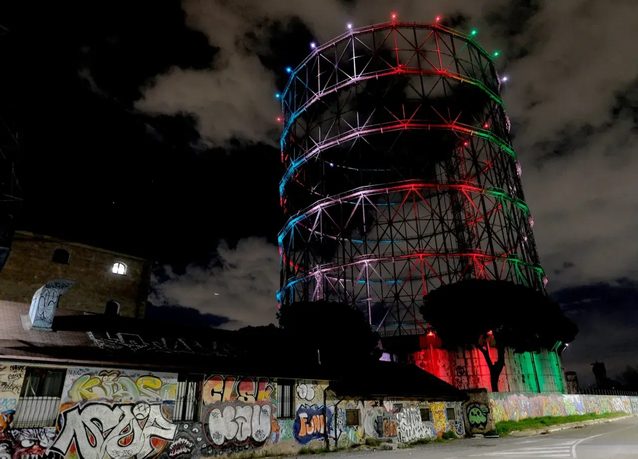 Il gasometro di Roma, illuminato da luci colorate, si staglia contro il cielo notturno. Un muro di graffiti decora l'edificio sottostante.