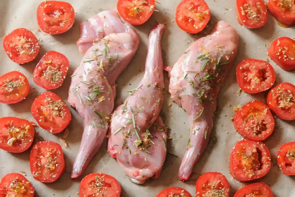 Trois gigolettes de lapin crues, assaisonnées d'herbes et d'ail, sont disposées avec des tomates coupées en deux, prêtes à être cuisinées.