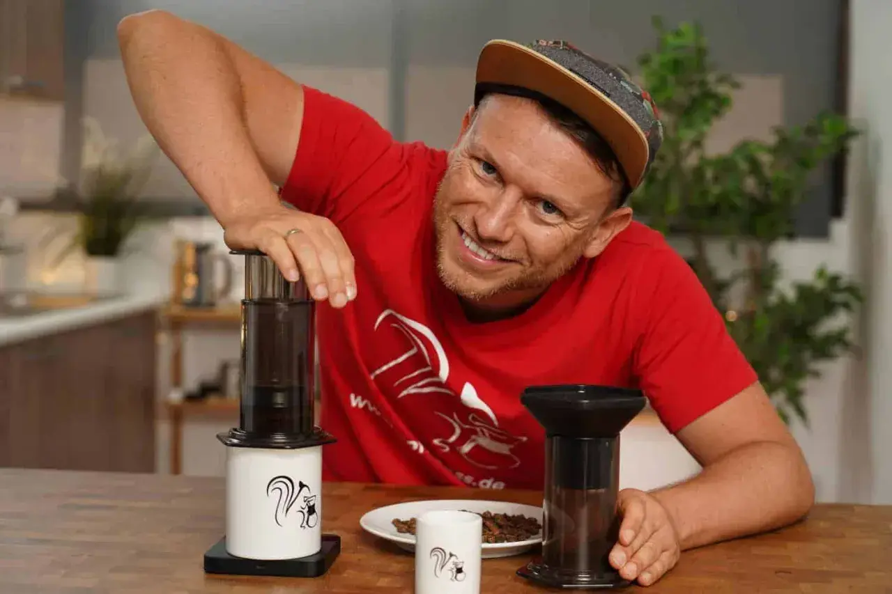 Mann mit rotem T-Shirt zeigt die Aeropress Zubereitung mit zwei Geräten und Kaffeebohnen.