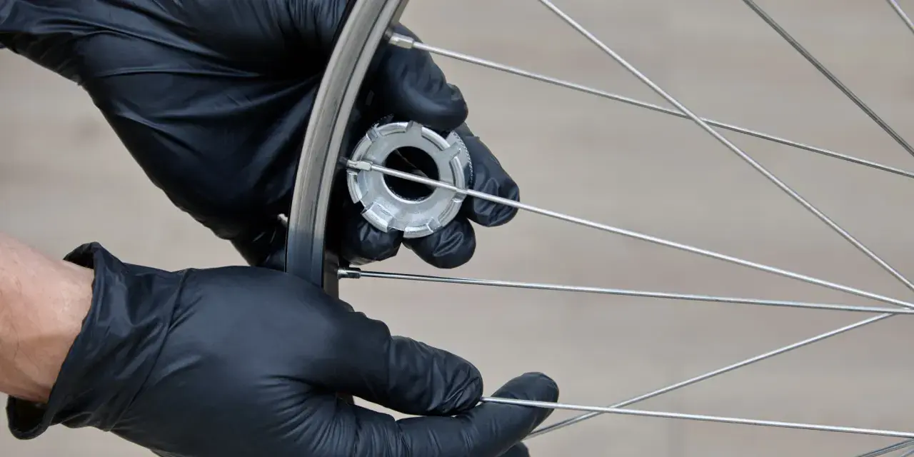 Hände mit schwarzen Handschuhen spannen die Speichen eines Fahrradrads mit einem Werkzeug.