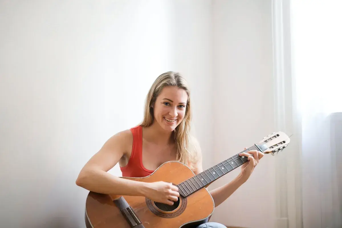 Lächelnde Frau mit blonden Haaren übt das Gitarre lernen für Erwachsene. Sie sitzt auf dem Boden und spielt auf einer Akustikgitarre.