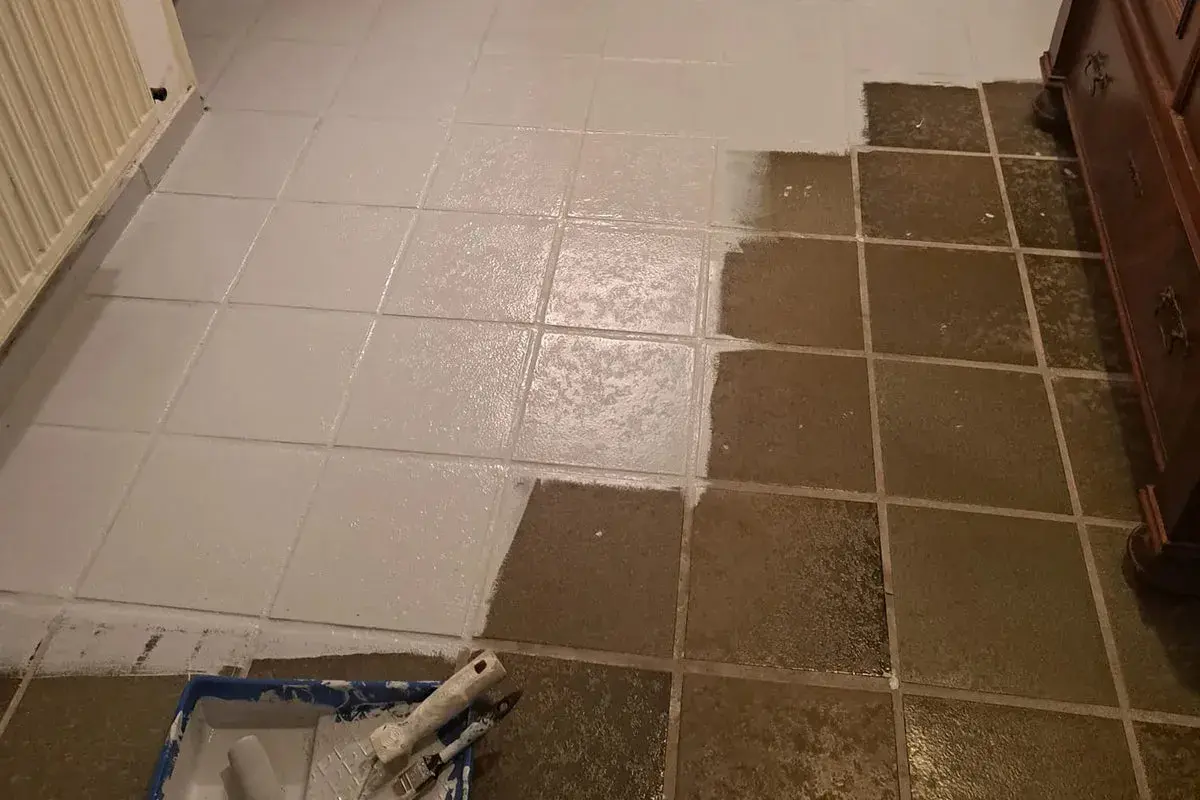 Bodenfliesen werden gerade weiß gestrichen. Ein Teil ist schon fertig, der andere noch dunkel. Werkzeug liegt bereit.