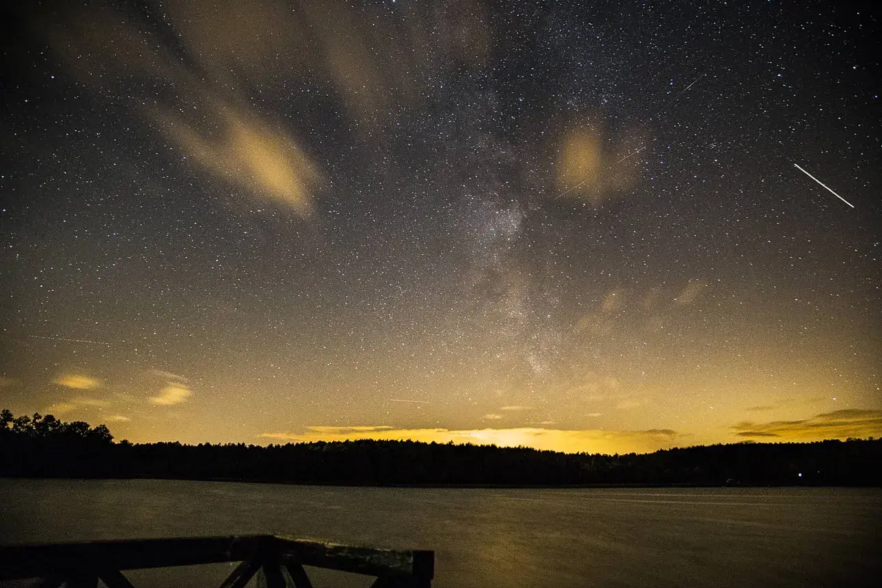 Nocne niebo nad jeziorem, pełne gwiazd i smug chmur, idealne do nauki, jak fotografować gwiazdy. Widać Drogę Mleczną i spadające gwiazdy.