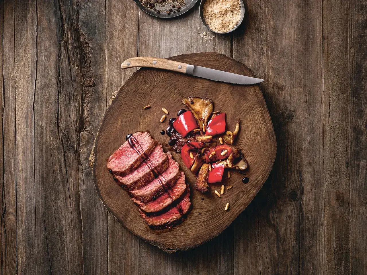 Saftige Tagliata di Manzo mit gebratenen Pilzen und Paprika auf einem rustikalen Holzbrett, garniert mit Pinienkernen und Balsamico.