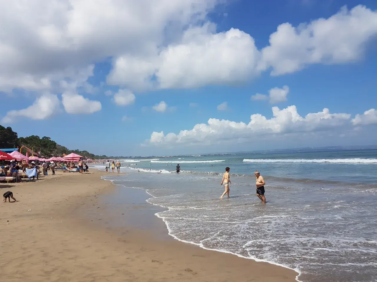 Słoneczny dzień na plaży w Seminyak na Bali. Ludzie spacerują po piasku i kąpią się w oceanie.