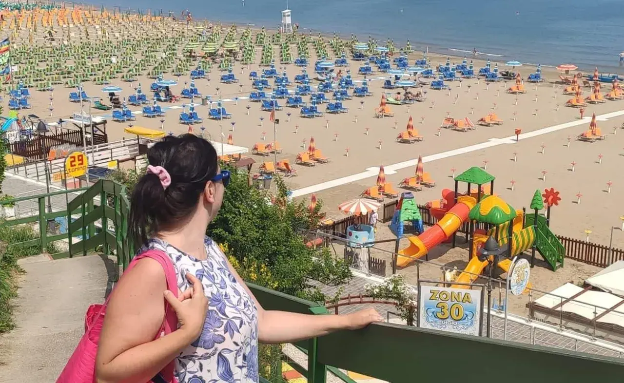 Donna con borsa rosa ammira la spiaggia di Gabicce Mare centro, con ombrelloni, lettini e un parco giochi colorato.