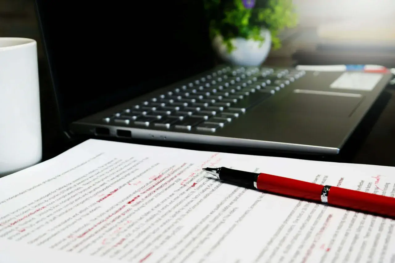 Czerwony długopis leży na dokumencie z poprawkami, obok laptopa. Widać tu różne odmiany cudzysłowu, które autor tekstu musiał poprawić.