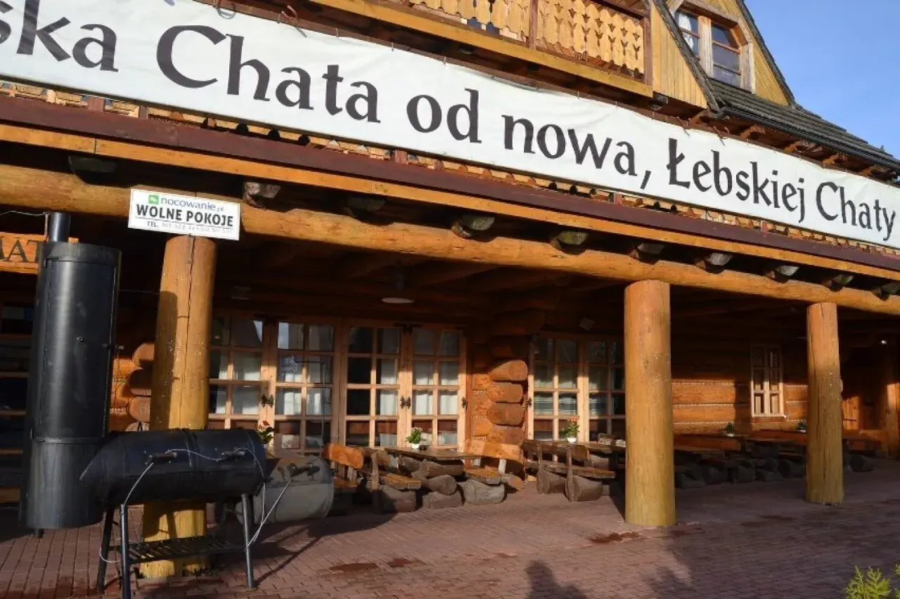 Drewniana, łebska chata z banerem "od nowa, Łebskiej Chaty". Przed wejściem grill i stoliki.