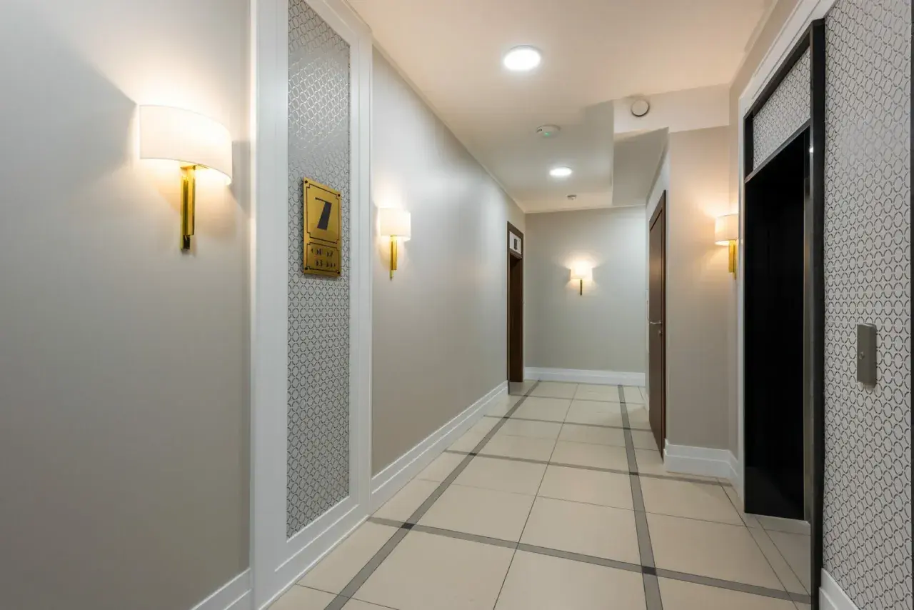 Korytarz hotelowy z jasnymi ścianami, co oznacza kolor szary, i eleganckimi lampami. Widać drzwi, windę i numer pokoju 7.